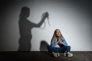Image showing Domestic violence, abusing. Scared little caucasian girl, victim sitting close to white wall with shadow of angry mother\'s threatening on it.