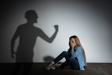 Image showing Domestic violence, abusing. Scared little caucasian girl, victim sitting close to white wall with shadow of angry father\'s threatening on it.