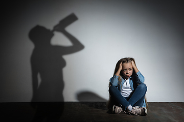 Image showing Domestic violence, abusing. Scared little caucasian girl, victim sitting close to white wall with shadow of angry threatening mother with alcohol addiction.