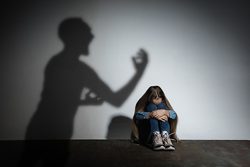 Image showing Domestic violence, abusing. Scared little caucasian girl, victim sitting close to white wall with shadow of angry father\'s threatening on it.