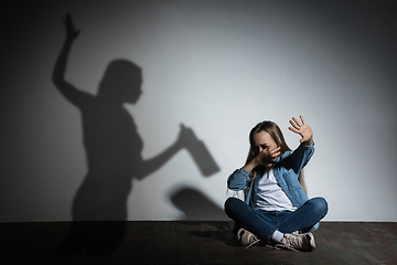 Image showing Domestic violence, abusing. Scared little caucasian girl, victim sitting close to white wall with shadow of angry threatening mother with alcohol addiction.