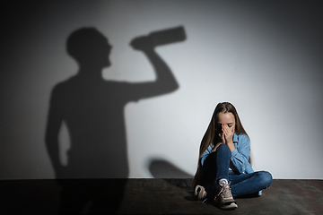 Image showing Domestic violence, abusing. Scared little caucasian girl, victim sitting close to white wall with shadow of angry threatening father with alcohol addiction.