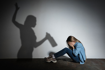 Image showing Domestic violence, abusing. Scared little caucasian girl, victim sitting close to white wall with shadow of angry threatening mother with alcohol addiction.