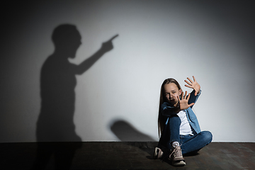 Image showing Domestic violence, abusing. Scared little caucasian girl, victim sitting close to white wall with shadow of angry father\'s threatening on it.