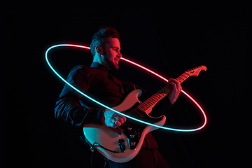 Image showing Young and joyful caucasian musician playing guitar on black studio background in neon light with glowing neoned circle. Concept of music, hobby, festival.