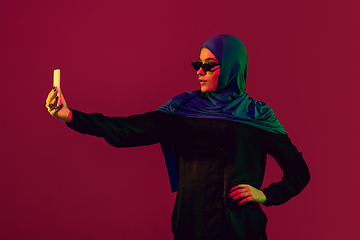 Image showing Beautiful arab woman posing in stylish hijab isolated on burgundy studio background in neon light. Fashion, beauty, style concept