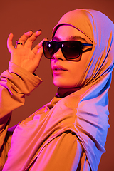 Image showing Beautiful arab woman posing in stylish hijab isolated on brown studio background in neon light. Fashion, beauty, style concept