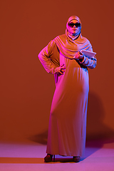 Image showing Beautiful arab woman posing in stylish hijab isolated on brown studio background in neon light. Fashion, beauty, style concept