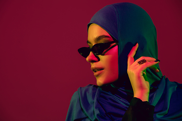 Image showing Beautiful arab woman posing in stylish hijab isolated on burgundy studio background in neon light. Fashion, beauty, style concept