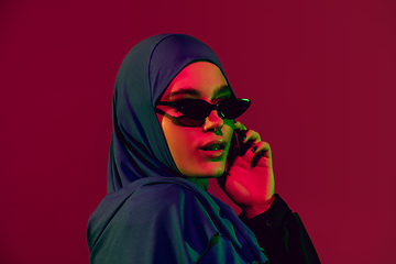 Image showing Beautiful arab woman posing in stylish hijab isolated on burgundy studio background in neon light. Fashion, beauty, style concept