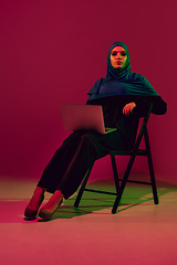 Image showing Beautiful arab woman posing in stylish hijab isolated on burgundy studio background in neon light. Fashion, beauty, style concept