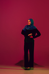 Image showing Beautiful arab woman posing in stylish hijab isolated on burgundy studio background in neon light. Fashion, beauty, style concept