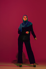 Image showing Beautiful arab woman posing in stylish hijab isolated on burgundy studio background in neon light. Fashion, beauty, style concept