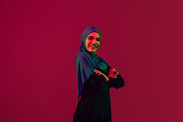 Image showing Beautiful arab woman posing in stylish hijab isolated on burgundy studio background in neon light. Fashion, beauty, style concept