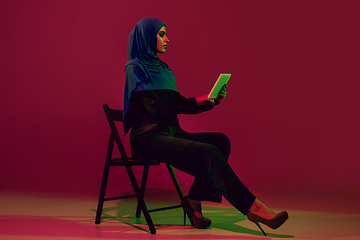 Image showing Beautiful arab woman posing in stylish hijab isolated on burgundy studio background in neon light. Fashion, beauty, style concept