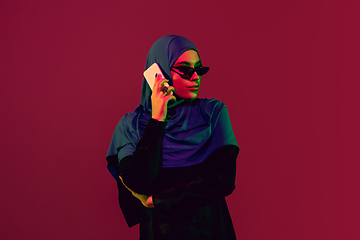 Image showing Beautiful arab woman posing in stylish hijab isolated on burgundy studio background in neon light. Fashion, beauty, style concept