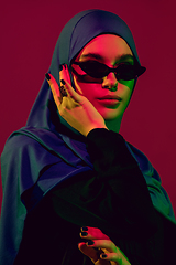 Image showing Beautiful arab woman posing in stylish hijab isolated on burgundy studio background in neon light. Fashion, beauty, style concept