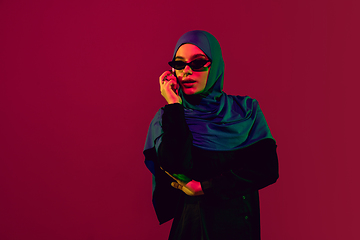 Image showing Beautiful arab woman posing in stylish hijab isolated on burgundy studio background in neon light. Fashion, beauty, style concept