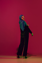 Image showing Beautiful arab woman posing in stylish hijab isolated on burgundy studio background in neon light. Fashion, beauty, style concept