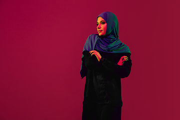 Image showing Beautiful arab woman posing in stylish hijab isolated on burgundy studio background in neon light. Fashion, beauty, style concept