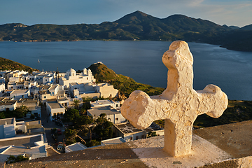 Image showing Christian cross and Plaka village on Milos island over red geranium flowers on sunset in Greece