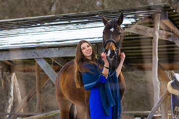 Image showing A girl in a blue dress hugs a horse against the backdrop of a snow-covered canopy in the forest