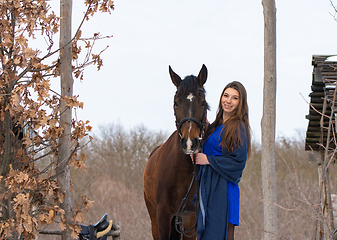 Image showing A girl in a blue dress and a horse on the background of a winter forest