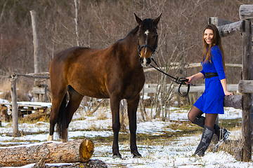 Image showing A beautiful girl in a short blue dress walks with a horse at an old farm in winter, the girl smiles and looks into the frame