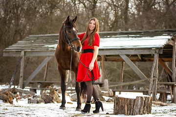 Image showing A beautiful girl in a red dress walks with a horse in the rays of the setting sun against the backdrop of old ruins