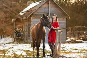 Image showing A beautiful girl in a red dress walks through an old farm in the winter in the rays of the setting sun