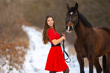 Image showing A beautiful girl in a red dress stands with a horse on the background of a winter road