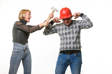 Image showing The wife screams and hits her husband on the head with a hammer, the husband is wearing a helmet and he covered his ears with his fingers