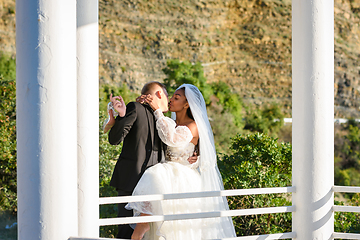 Image showing Young beautiful interracial newlyweds are kissing in the gazebo, against the backdrop of sunny mountains