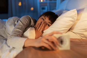 Image showing sleepy asian woman with clock in bed at night