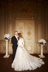 Image showing Luxury wedding couple in love. Beautiful bride in white dress with brides bouquet and handsome groom in black suit standing in baroque interior and embracing each other