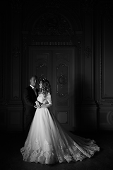 Image showing Black and white photography. Luxury wedding couple in love. Beautiful bride in white dress with brides bouquet and handsome groom in black suit standing in baroque interior and embracing each other