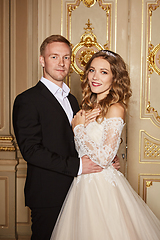 Image showing Luxury wedding couple in love. Beautiful bride in white dress with brides bouquet and handsome groom in black suit standing in baroque interior and embracing each other