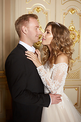 Image showing Luxury wedding couple in love. Beautiful bride in white dress with brides bouquet and handsome groom in black suit standing in baroque interior and embracing each other