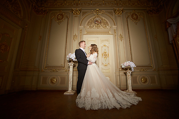 Image showing Luxury wedding couple in love. Beautiful bride in white dress with brides bouquet and handsome groom in black suit standing in baroque interior and embracing each other