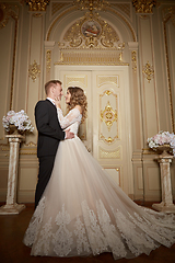 Image showing Luxury wedding couple in love. Beautiful bride in white dress with brides bouquet and handsome groom in black suit standing in baroque interior and embracing each other