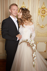Image showing Luxury wedding couple in love. Beautiful bride in white dress with brides bouquet and handsome groom in black suit standing in baroque interior and embracing each other