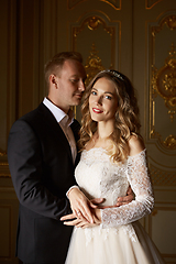 Image showing Luxury wedding couple in love. Beautiful bride in white dress with brides bouquet and handsome groom in black suit standing in baroque interior and embracing each other