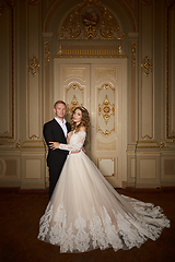 Image showing Luxury wedding couple in love. Beautiful bride in white dress with brides bouquet and handsome groom in black suit standing in baroque interior and embracing each other