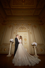 Image showing Luxury wedding couple in love. Beautiful bride in white dress with brides bouquet and handsome groom in black suit standing in baroque interior and embracing each other