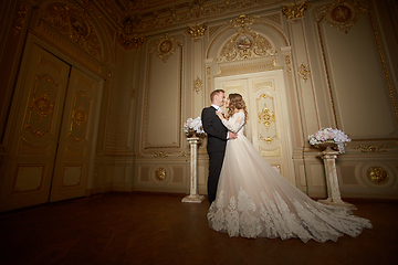 Image showing Luxury wedding couple in love. Beautiful bride in white dress with brides bouquet and handsome groom in black suit standing in baroque interior and embracing each other