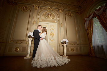 Image showing Luxury wedding couple in love. Beautiful bride in white dress with brides bouquet and handsome groom in black suit standing in baroque interior and embracing each other