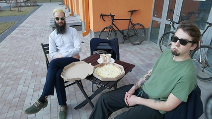 Image showing Friendly talk. Two happy young men eating pizza and talking to each other while sitting outdoors.