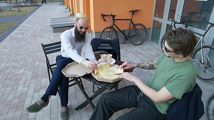 Image showing Friendly talk. Two happy young men eating pizza and talking to each other while sitting outdoors.