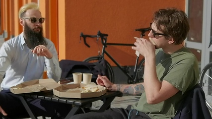 Image showing Friendly talk. Two happy young men eating pizza and talking to each other while sitting outdoors