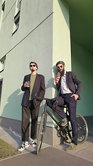 Image showing Two young men friends dressed casually spending time together at the city, drinking takeaway coffee next bike.